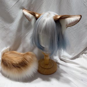 Puede incluir: Un conjunto que incluye una peluca corta y recta de color azul claro con flequillo, adornada con grandes orejas de animal puntiagudas de piel sintética marrón y blanca. Al lado hay una cola a juego de piel sintética gruesa.