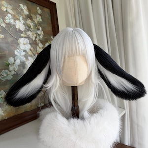 以下が含まれることがあります： 白と黒のウィッグ、長くふわふわのウサギの耳付き。ウィッグは白いベースに耳の黒いアクセントがあります。白い毛皮の襟がウィッグの根元にあります。ウィッグはマネキンの頭に展示されています。