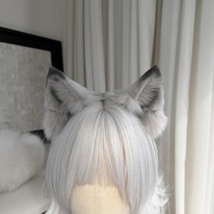 Puede incluir: Un accesorio de cosplay con una peluca de pelo largo plateado y un par de orejas de lobo grises y blancas. Las orejas son esponjosas y detalladas, con puntas grises m&aacute;s oscuras. La peluca tiene flequillo.