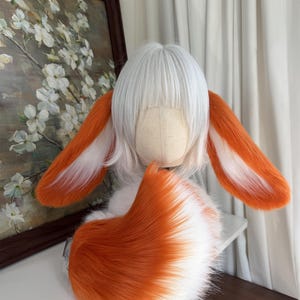 May include: A white wig with straight bangs styled on a mannequin head, featuring two long, plush, orange and white faux fur ears attached to the sides and a large, matching orange and white faux fur tail curled at the base of the head.