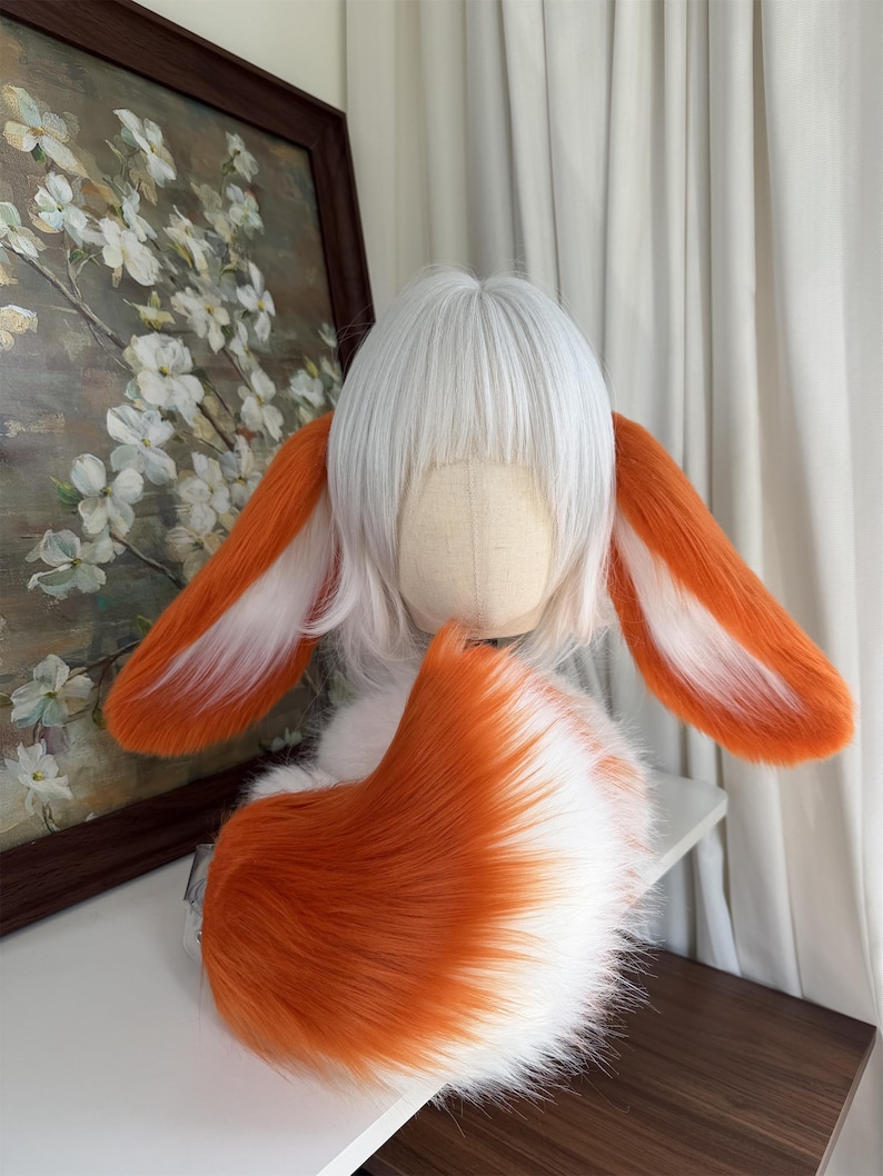 May include: A white wig with straight bangs styled on a mannequin head, featuring two long, plush, orange and white faux fur ears attached to the sides and a large, matching orange and white faux fur tail curled at the base of the head.