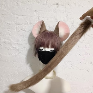 May include: A set of faux fur mouse ears and a long matching tail. The ears are brown with pink inner sections, attached to a wig with short mauve hair. A black face mask covers the lower face, and a white beaded necklace rests on the neck.