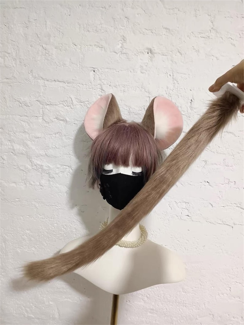 May include: A set of faux fur mouse ears and a long matching tail. The ears are brown with pink inner sections, attached to a wig with short mauve hair. A black face mask covers the lower face, and a white beaded necklace rests on the neck.