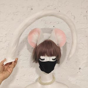 May include: A white faux fur tail accessory and a pair of plush mouse ears with pink inner detailing. The ears are positioned on a mannequin head featuring short brown hair, a black face mask, and a multi-strand white pearl necklace.