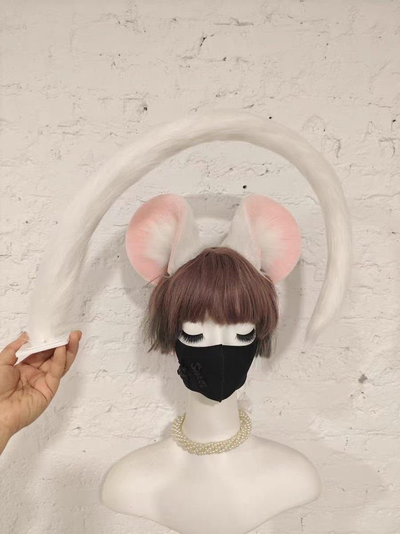 May include: A white faux fur tail accessory and a pair of plush mouse ears with pink inner detailing. The ears are positioned on a mannequin head featuring short brown hair, a black face mask, and a multi-strand white pearl necklace.