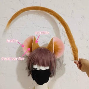 May include: A mannequin head wearing a short purple-toned wig, a black face mask, and a pearl necklace. Attached are large, rounded animal ears with light pink inner surfaces and golden-brown outer fur. A long, thin, golden-brown tail is held up.