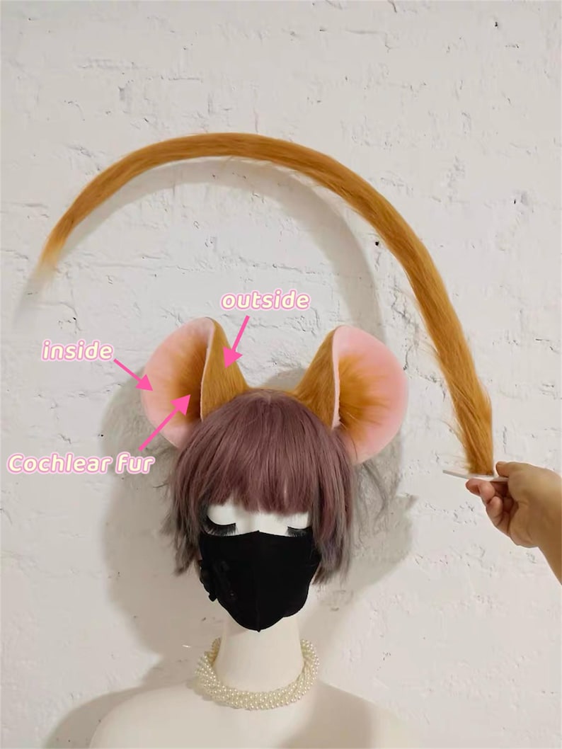 May include: A mannequin head wearing a short purple-toned wig, a black face mask, and a pearl necklace. Attached are large, rounded animal ears with light pink inner surfaces and golden-brown outer fur. A long, thin, golden-brown tail is held up.