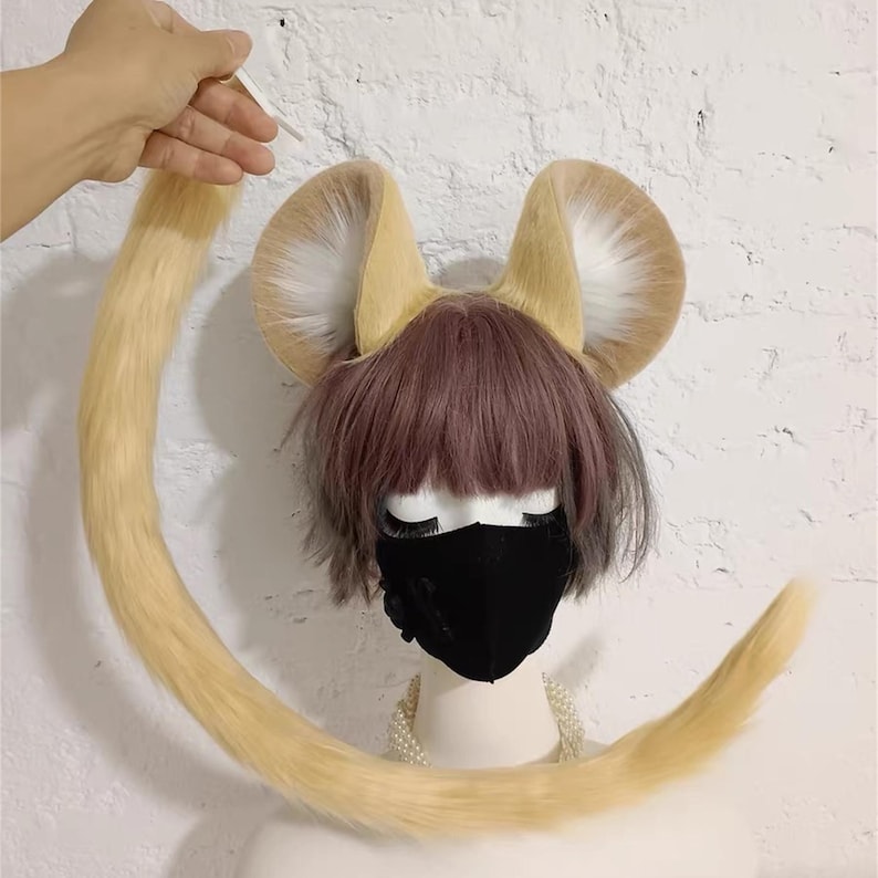 May include: A set of light tan and white faux fur animal ears attached to a headband, paired with a long, matching light tan faux fur tail. The ears feature a rounded shape with white inner fur and tan outer fur, displayed on a mannequin head.
