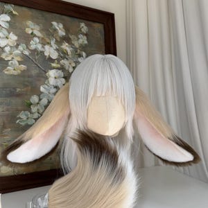 May include: A white wig with straight bangs and long layers, featuring large, plush, animal-like ears attached to the sides. The ears are tan with dark brown tips and pinkish-white inner details. A matching, thick, multi-toned faux fur tail is included.