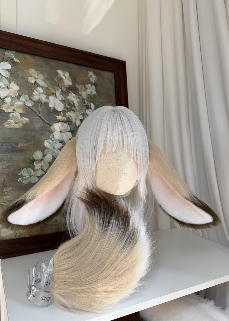 May include: A white wig with straight bangs and long layers, featuring large, plush, animal-like ears attached to the sides. The ears are tan with dark brown tips and pinkish-white inner details. A matching, thick, multi-toned faux fur tail is included.