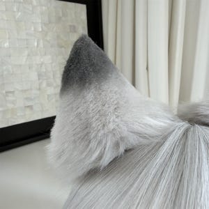 Puede incluir: Primer plano de una oreja de lobo de piel sint&eacute;tica gris y blanca. La oreja es de color gris claro con una punta gris m&aacute;s oscura. La piel es larga y esponjosa, con una textura realista. La oreja es parte de un disfraz o accesorio.