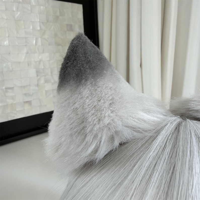 Puede incluir: Primer plano de una oreja de lobo de piel sint&eacute;tica gris y blanca. La oreja es de color gris claro con una punta gris m&aacute;s oscura. La piel es larga y esponjosa, con una textura realista. La oreja es parte de un disfraz o accesorio.