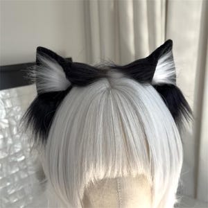 Op de afbeelding: Zwart en witte kattenoren hoofdband. De oren zijn zwart met witte vacht accenten. De hoofdband wordt gedragen op een hoofd met wit haar.