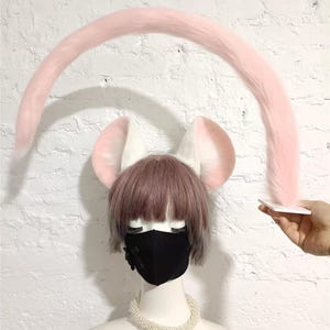 May include: A long, curved, light pink faux fur tail held by a white square base, positioned above a mannequin head wearing white animal ears with pink inner detailing, a short brown wig, a black face mask, and a white beaded necklace.