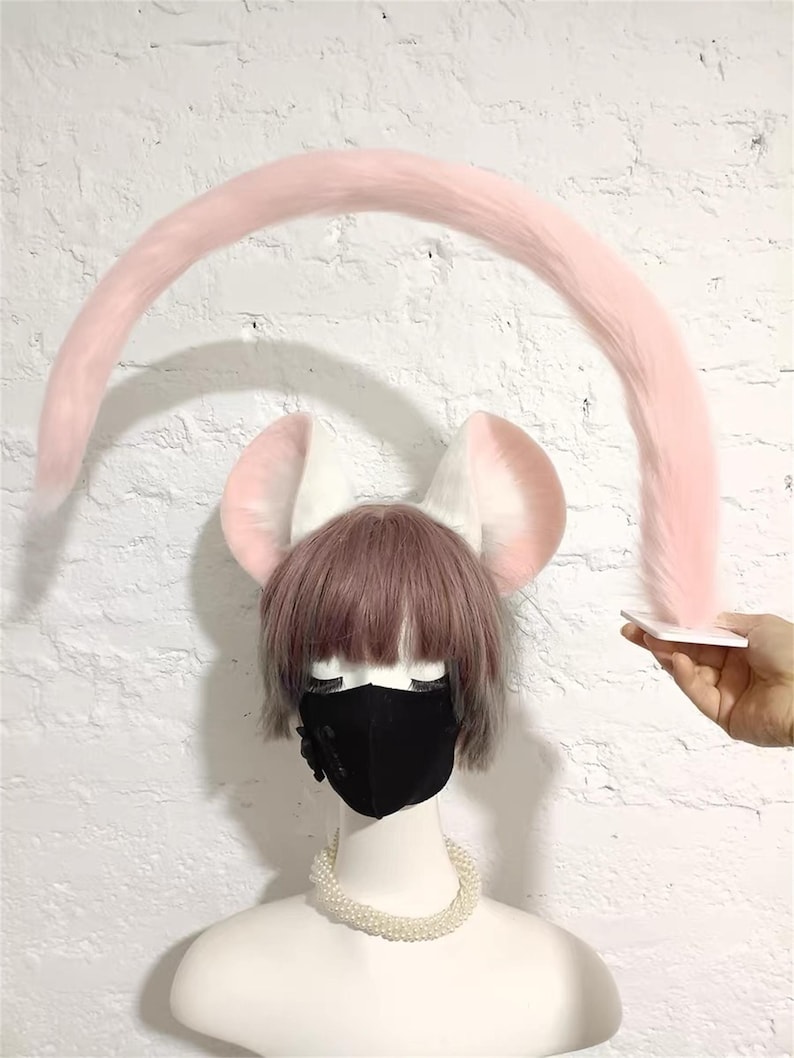 May include: A long, curved, light pink faux fur tail held by a white square base, positioned above a mannequin head wearing white animal ears with pink inner detailing, a short brown wig, a black face mask, and a white beaded necklace.