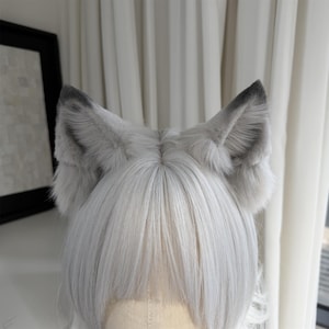 Puede incluir: Un par de orejas de lobo de piel sint&eacute;tica gris unidas a una diadema. Las orejas son esponjosas con puntas grises m&aacute;s oscuras y est&aacute;n colocadas sobre una peluca de pelo plateado. El fondo es una cortina blanca.