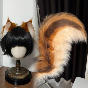 May include: A cosplay set featuring a black wig on a mannequin head with attached brown and white fox ears. A large, fluffy fox tail in shades of brown, orange, and white is displayed next to the wig.