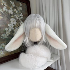 May include: A white wig with straight bangs and shoulder-length hair, featuring two large, plush, white rabbit ears with soft pink inner detailing. A fluffy white faux fur collar or accessory is positioned at the base of the wig head form.