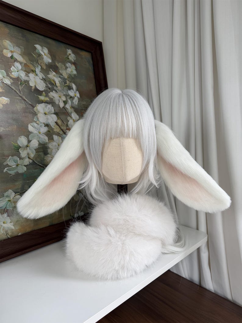 May include: A white wig with straight bangs and shoulder-length hair, featuring two large, plush, white rabbit ears with soft pink inner detailing. A fluffy white faux fur collar or accessory is positioned at the base of the wig head form.