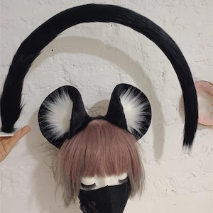May include: A set of black and white faux fur animal ears on a headband and a long, curved black faux fur tail. The ears feature black outer edges with white inner fur. The tail is arched, and the ears are positioned on a mannequin head.