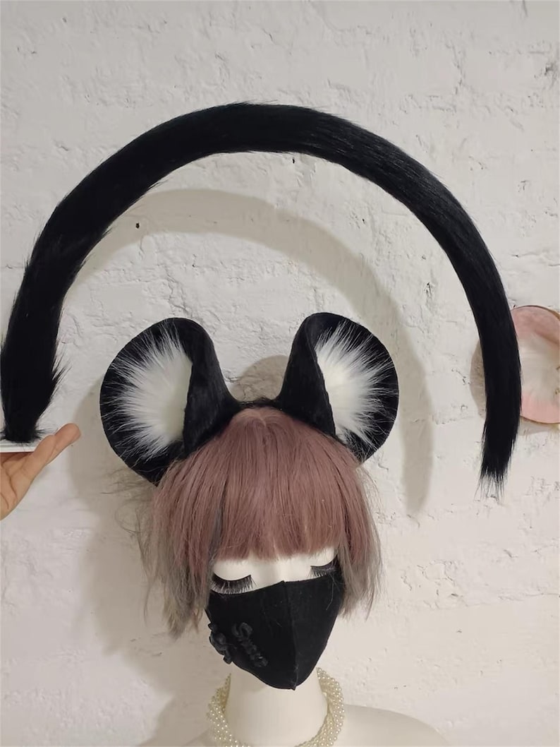 May include: A set of black and white faux fur animal ears on a headband and a long, curved black faux fur tail. The ears feature black outer edges with white inner fur. The tail is arched, and the ears are positioned on a mannequin head.