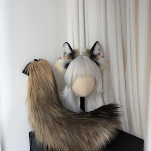 May include: A faux fur fox tail and ear set. The tail is a gradient of brown and tan, with a black ribbon. The ears are black, white, and tan. The set is displayed on a black surface against a white backdrop.