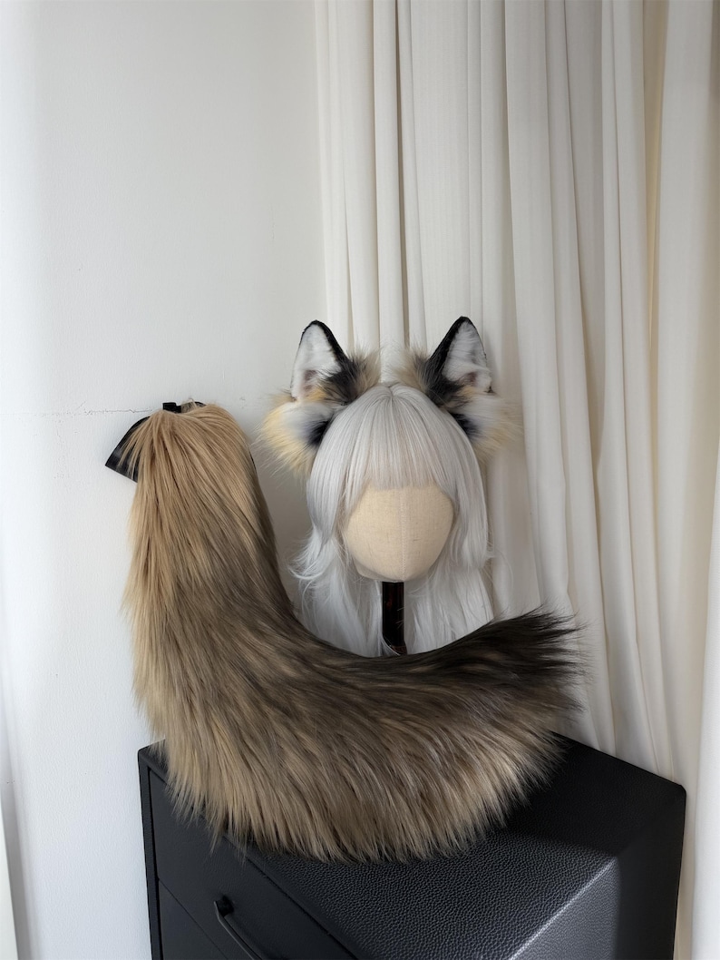 May include: A faux fur fox tail and ear set. The tail is a gradient of brown and tan, with a black ribbon. The ears are black, white, and tan. The set is displayed on a black surface against a white backdrop.