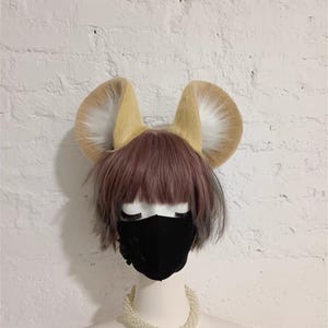 May include: A pair of large, upright, tan and white faux fur animal ears attached to a headband. The ears are positioned on a mannequin head featuring a short, layered, dusty pink wig and a solid black face mask, accented by a white pearl necklace.