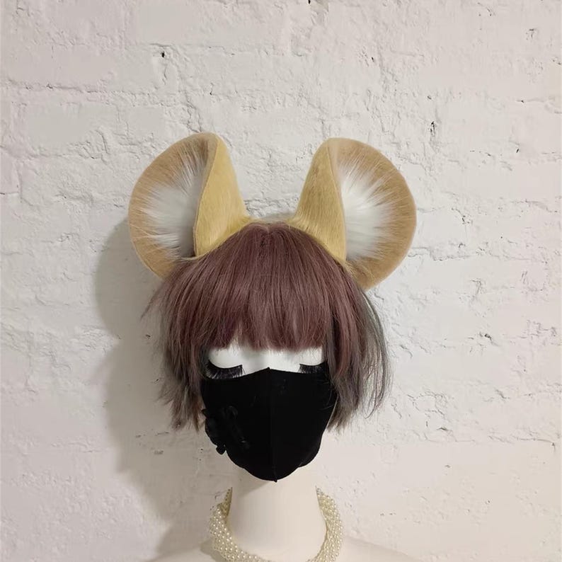 May include: A pair of large, upright, tan and white faux fur animal ears attached to a headband. The ears are positioned on a mannequin head featuring a short, layered, dusty pink wig and a solid black face mask, accented by a white pearl necklace.