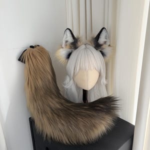 May include: A faux fur fox tail and ear set. The tail is a blend of brown and tan, with a black attachment. The ears are black, white, and tan. The ears are attached to a wig head with white hair.