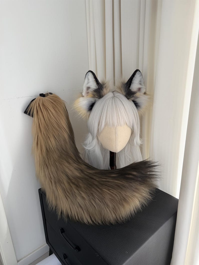 May include: A faux fur fox tail and ear set. The tail is a blend of brown and tan, with a black attachment. The ears are black, white, and tan. The ears are attached to a wig head with white hair.