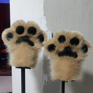 May include: A pair of tan faux fur animal paw gloves featuring four rounded black pads and a larger central black palm pad on each hand. The plush material covers the entire exterior, creating a soft, textured appearance for these hand coverings.