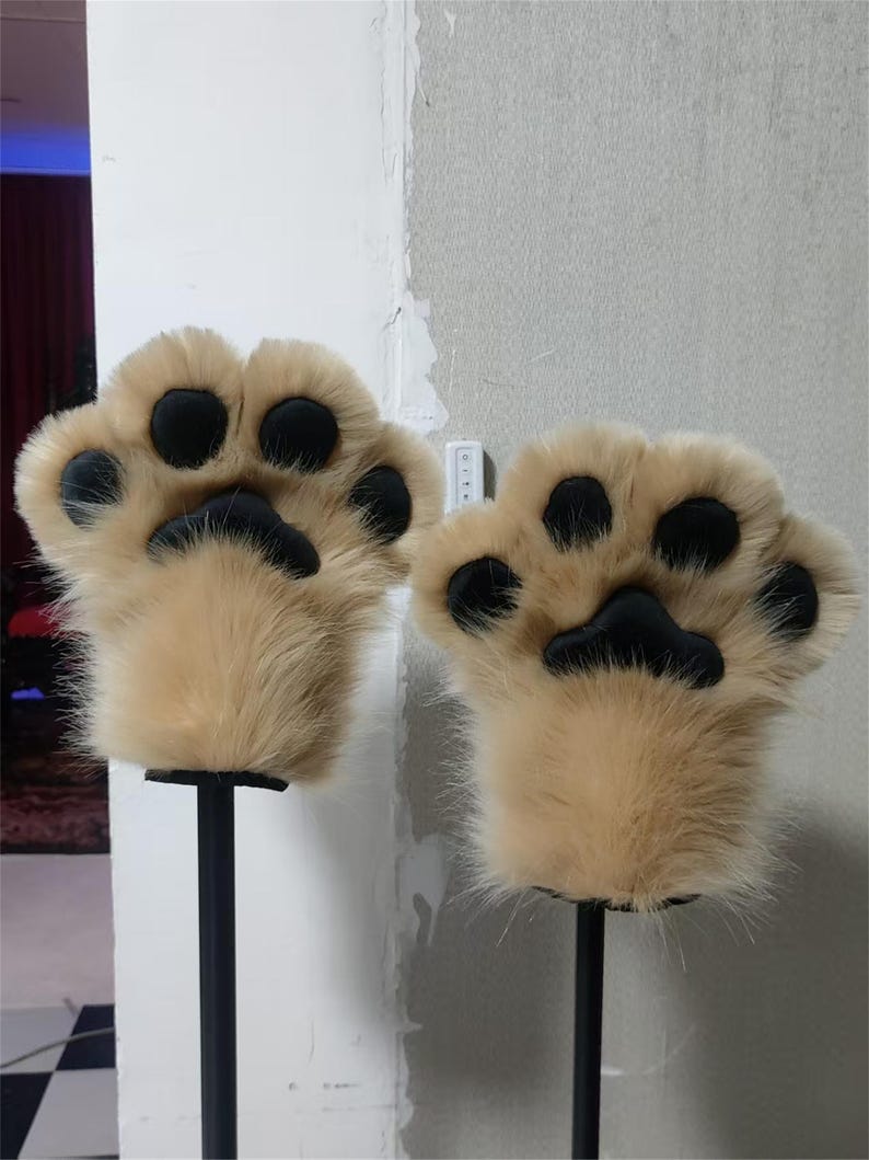 May include: A pair of tan faux fur animal paw gloves featuring four rounded black pads and a larger central black palm pad on each hand. The plush material covers the entire exterior, creating a soft, textured appearance for these hand coverings.