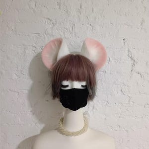 May include: A mannequin head wearing a short brown wig with bangs, a black face mask, and a white pearl necklace. Attached to the top of the head are two large, rounded, plush animal ears that are white with soft pink centers and edges.