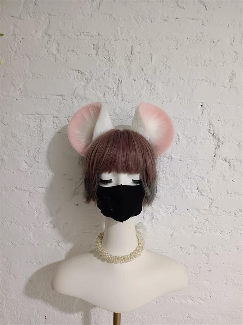 May include: A mannequin head wearing a short brown wig with bangs, a black face mask, and a white pearl necklace. Attached to the top of the head are two large, rounded, plush animal ears that are white with soft pink centers and edges.