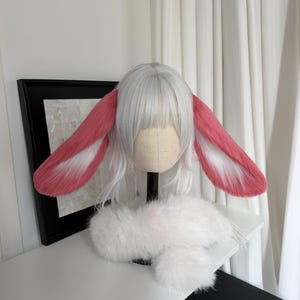 May include: A white wig with straight bangs and long side strands, featuring large, plush, pink and white rabbit ears attached to the sides. A fluffy white faux fur collar or neck piece is positioned at the base of the wig on a mannequin head.