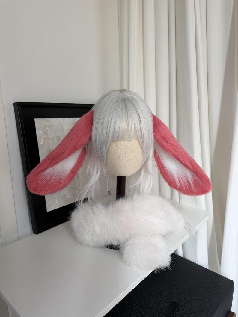 May include: A white wig with straight bangs and long side strands, featuring large, plush, pink and white rabbit ears attached to the sides. A fluffy white faux fur collar or neck piece is positioned at the base of the wig on a mannequin head.