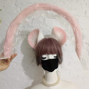May include: A set of plush animal ears in white and soft pink, paired with a long, curved, light pink tail encased in clear plastic packaging. The ears are positioned atop a mannequin head featuring a short, dusty rose wig and a black face mask.