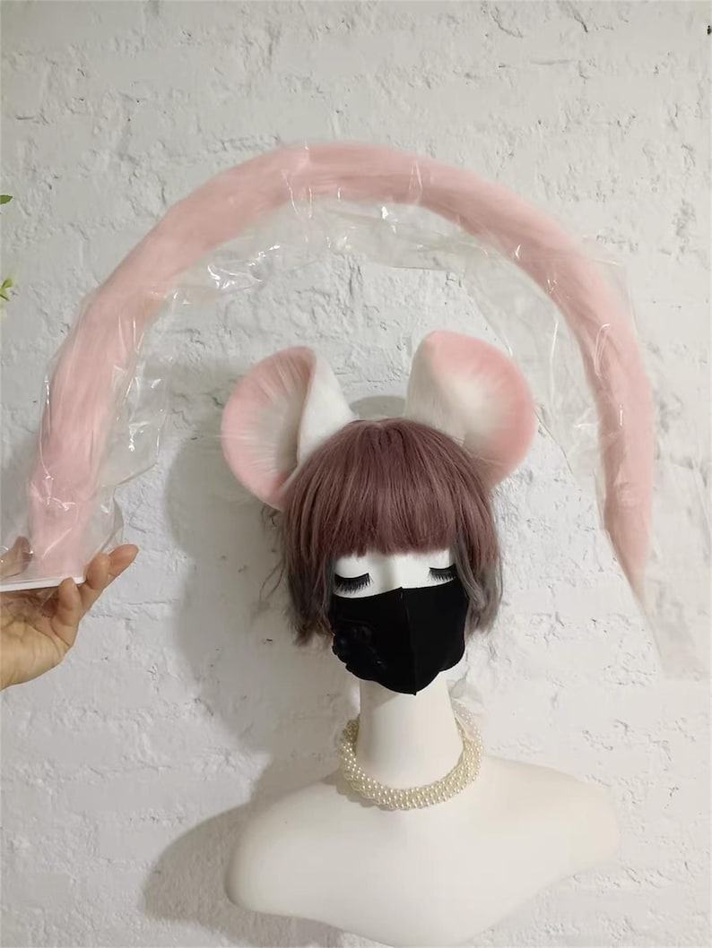 May include: A set of plush animal ears in white and soft pink, paired with a long, curved, light pink tail encased in clear plastic packaging. The ears are positioned atop a mannequin head featuring a short, dusty rose wig and a black face mask.
