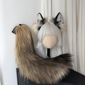 May include: A faux fur fox tail and ear set. The tail is a blend of brown and tan, with a black ribbon at the top. The ears are white, black, and tan, attached to a headband. The set is designed for cosplay or costume use.