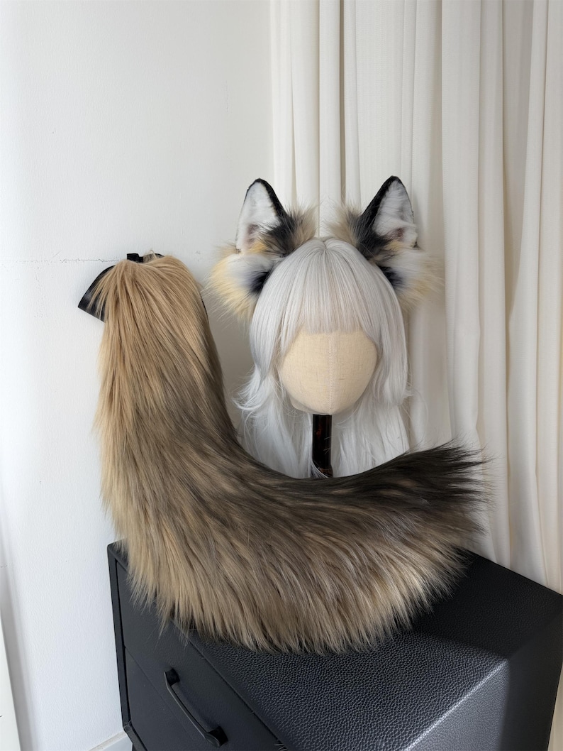 May include: A faux fur fox tail and ear set. The tail is a blend of brown and tan, with a black ribbon at the top. The ears are white, black, and tan, attached to a headband. The set is designed for cosplay or costume use.