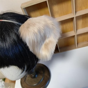 May include: A headband featuring a pair of plush, faux fur animal ears in a light beige and cream color palette. The soft, textured ears are attached to a thin, flexible band, designed to sit atop the head with a natural, upright appearance.