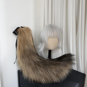 May include: A long, faux fur tail in shades of brown and tan, with a black ribbon at the top. The tail is displayed on a black surface, with a white wig on a mannequin head in the background.
