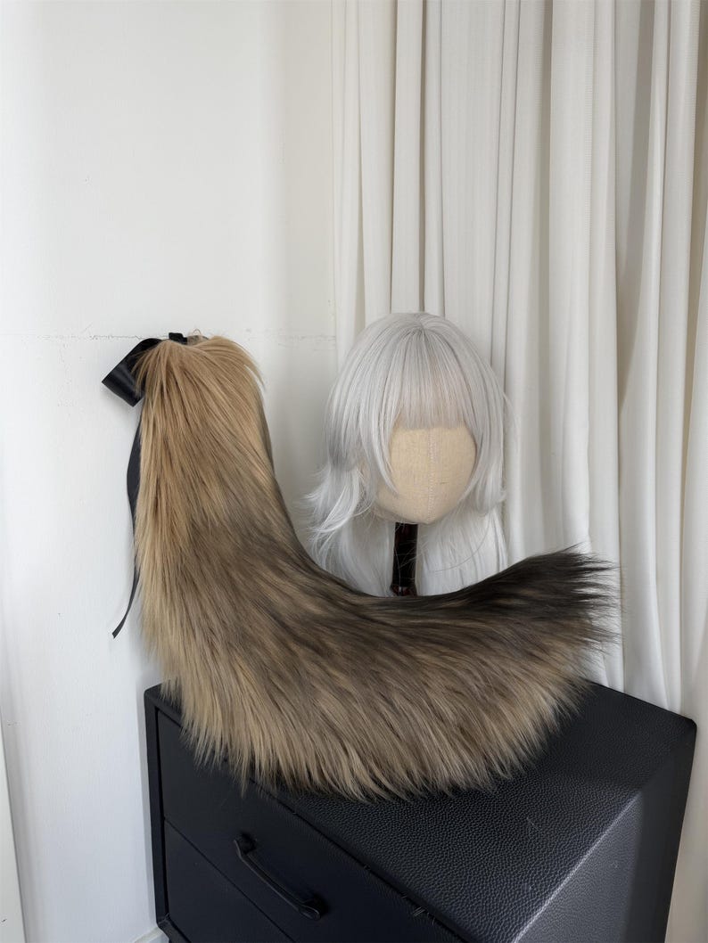 May include: A long, faux fur tail in shades of brown and tan, with a black ribbon at the top. The tail is displayed on a black surface, with a white wig on a mannequin head in the background.