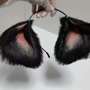May include: A black headband featuring two large, pointed animal ears covered in long, black faux fur. The inner portion of each ear is lined with a soft, light pink fabric, and the ears are attached to a thin, flexible black metal band.