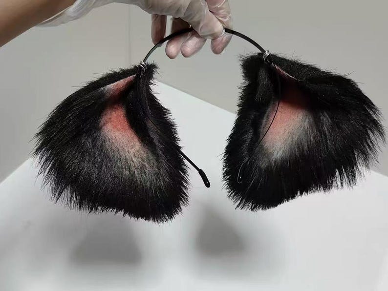May include: A black headband featuring two large, pointed animal ears covered in long, black faux fur. The inner portion of each ear is lined with a soft, light pink fabric, and the ears are attached to a thin, flexible black metal band.
