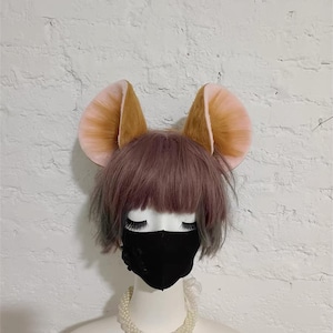 May include: A mannequin head wearing a short, dusty purple wig, a black face mask, and a white pearl necklace. Attached to the top of the head are two large, rounded animal ears with brown fur on the outer edges and light pink inner ear details.