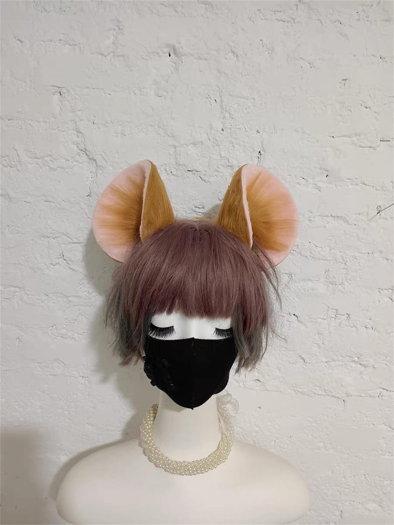 May include: A mannequin head wearing a short, dusty purple wig, a black face mask, and a white pearl necklace. Attached to the top of the head are two large, rounded animal ears with brown fur on the outer edges and light pink inner ear details.