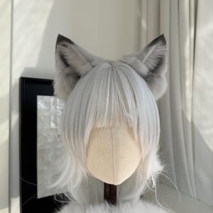 Puede incluir: Peluca gris y blanca con orejas de lobo de piel sint&eacute;tica adjuntas. La peluca tiene pelo largo, liso y blanco con flequillo. Las orejas son grises con puntas grises m&aacute;s oscuras. La peluca se exhibe en una cabeza de maniqu&iacute;.
