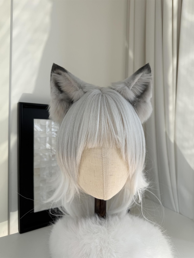 Puede incluir: Peluca gris y blanca con orejas de lobo de piel sint&eacute;tica adjuntas. La peluca tiene pelo largo, liso y blanco con flequillo. Las orejas son grises con puntas grises m&aacute;s oscuras. La peluca se exhibe en una cabeza de maniqu&iacute;.